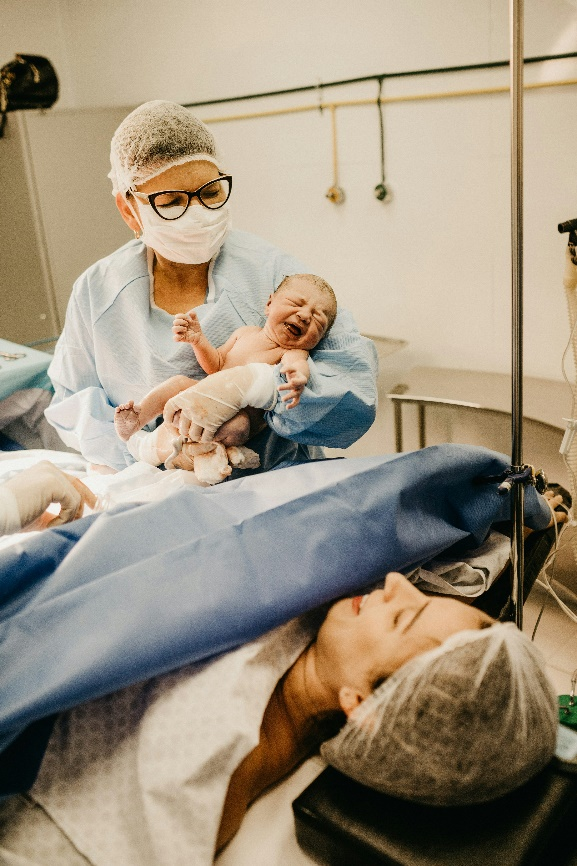 A healthcare professional in surgical attire holding a crying newborn baby while the mother lies in the hospital bed after delivery.